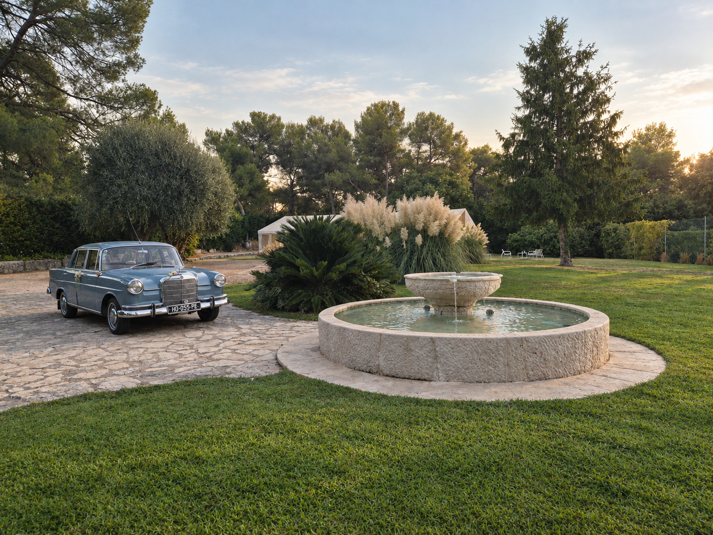 Voiture ancienne bleu clair garée sur le patio de pierre face à la fontaine du Mas Baïeta au coucher du soleil
